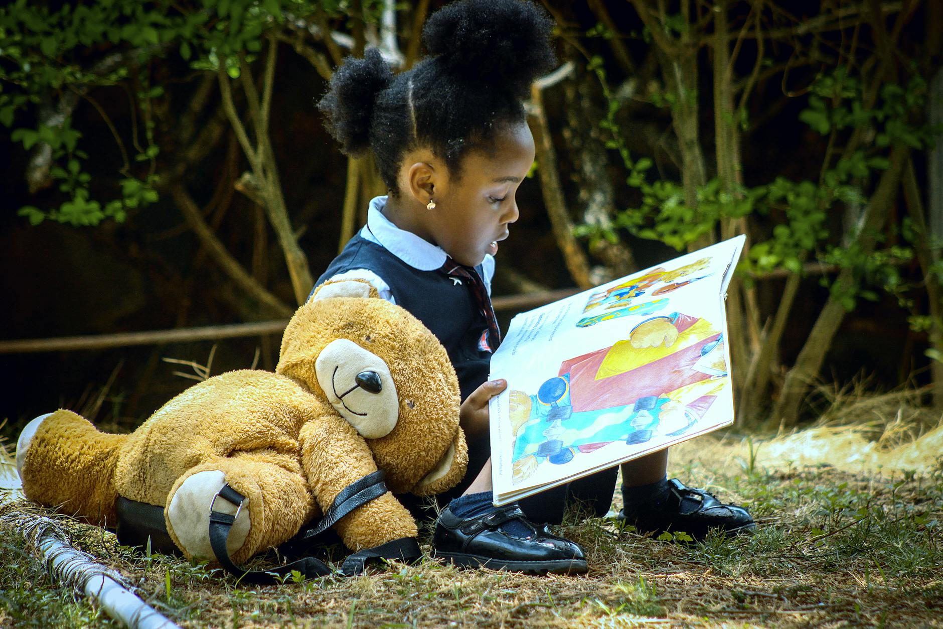 a little girl sitting on the grass with a teddy bear and a book