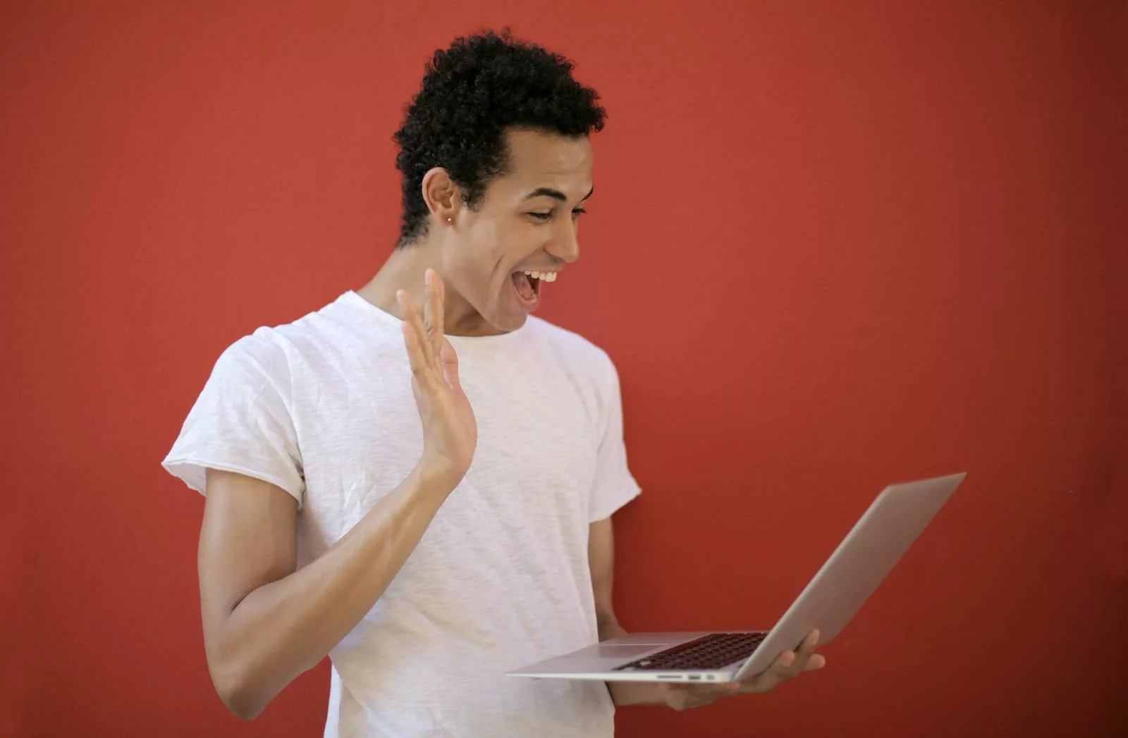 Young man making a cheerful video call on a laptop with a red background, expressing joy and surprise.