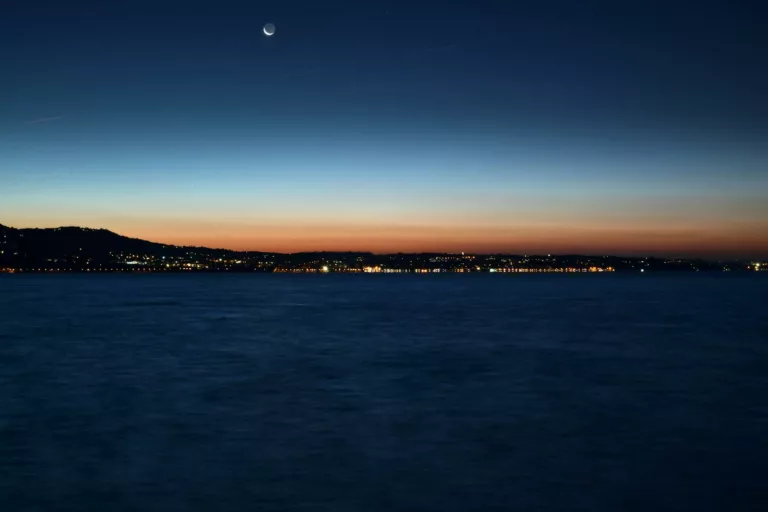 A tranquil view of Garda Lake under a crescent moon at twilight, capturing serene water and distant city lights.