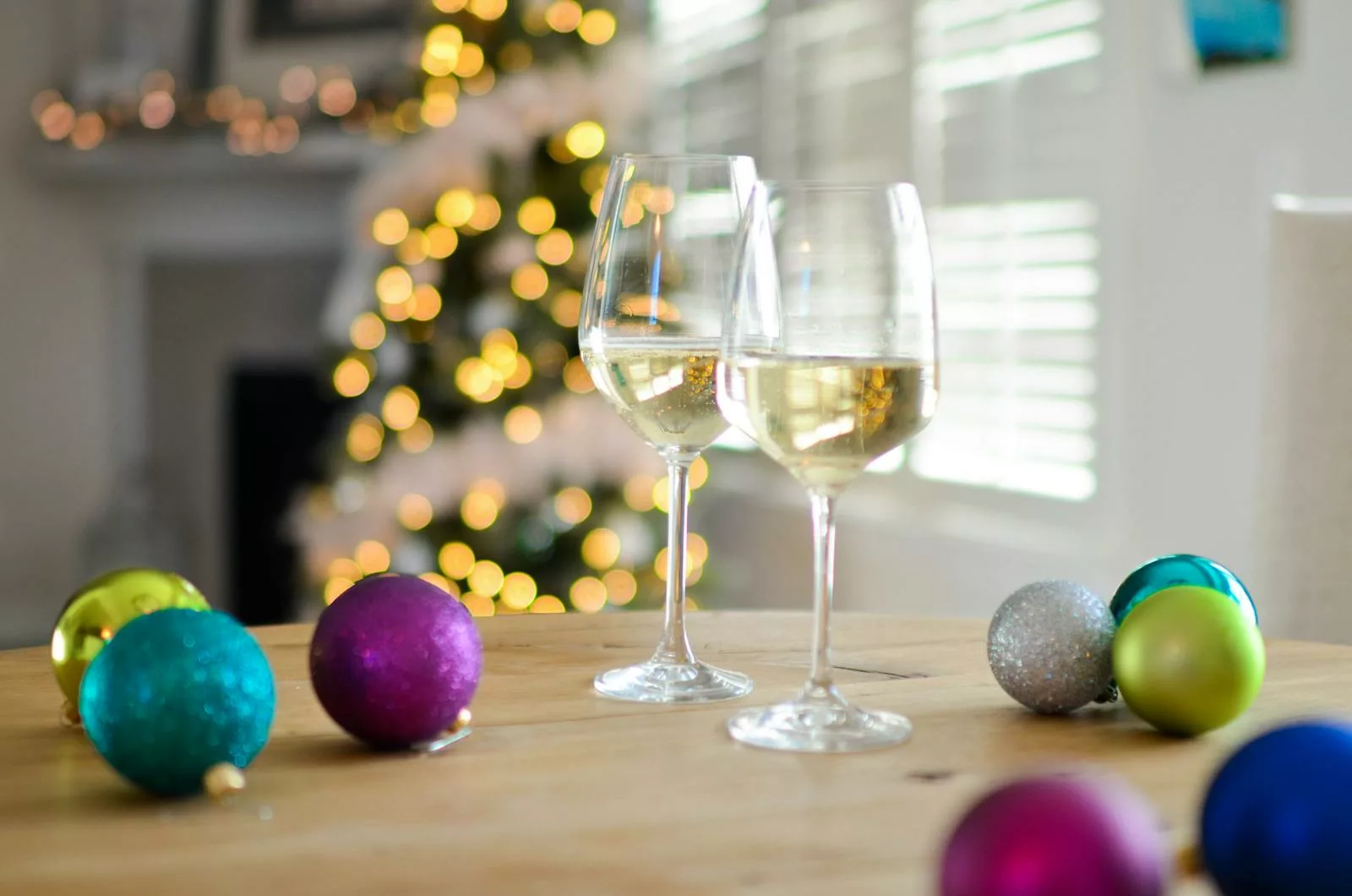 Two glasses of white wine on a table with colorful Christmas baubles and blurred tree lights.