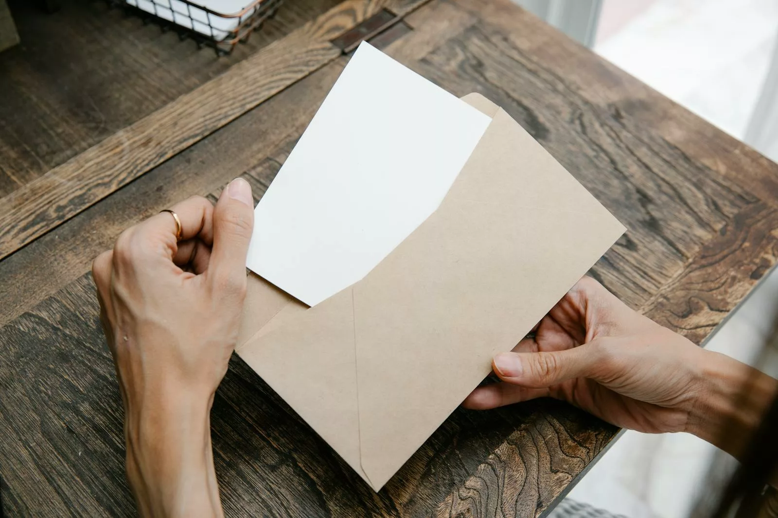 Hands holding a letter inside a brown envelope on a wooden desk, viewed from above.