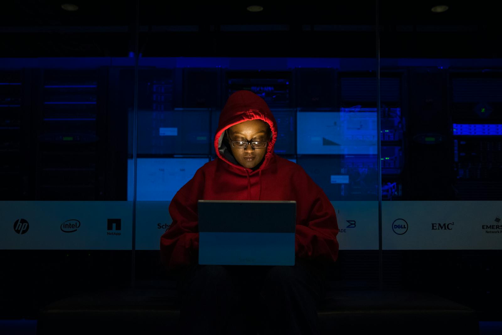 Focused woman in a red hoodie using a laptop in a dimly lit room with technology brands displayed.