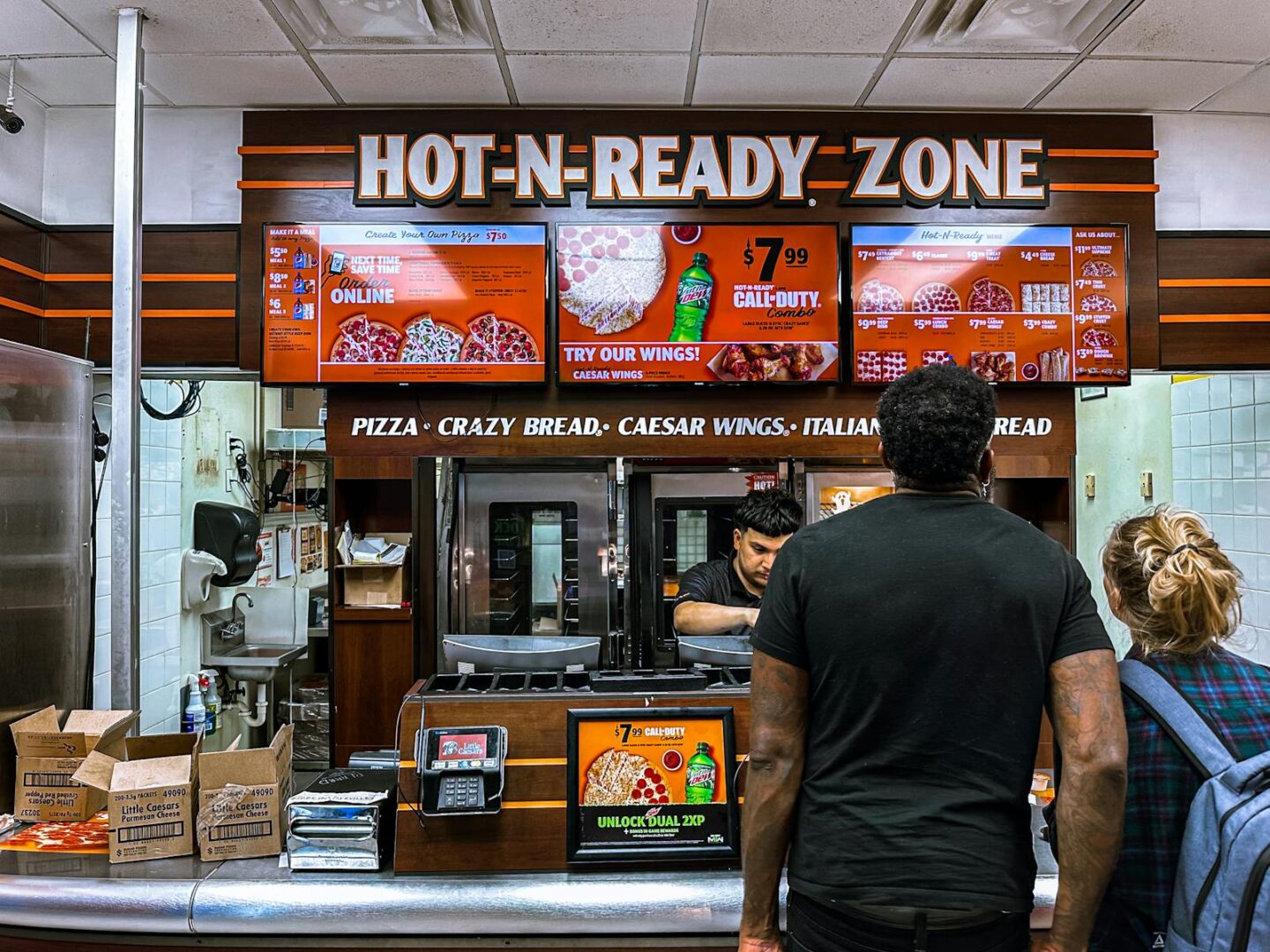 Busy fast food restaurant with customers ordering pizza at the counter under bright digital menu signage.