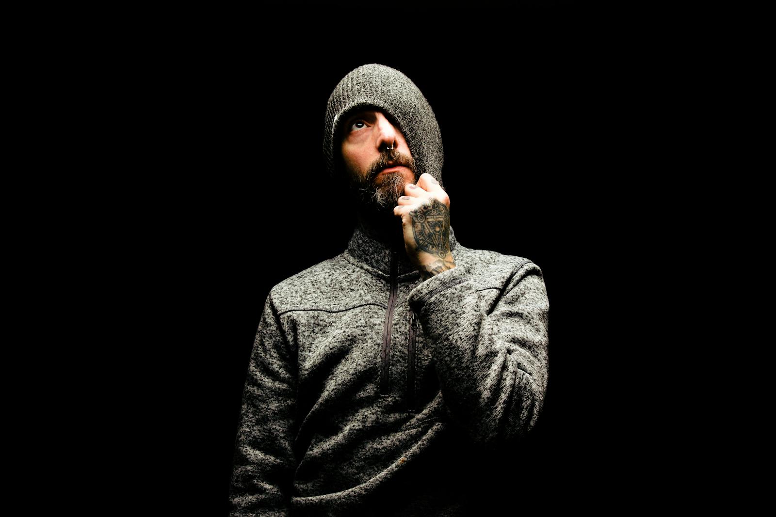 Close-up of a bearded man in a sweater and beanie, deep in thought against a dark background.