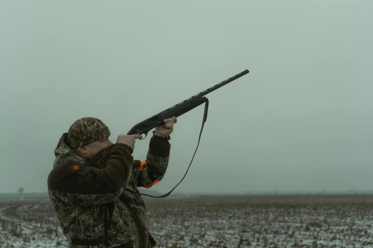 A hunter in camouflage aims a rifle in a foggy open field, showcasing outdoor hunting.